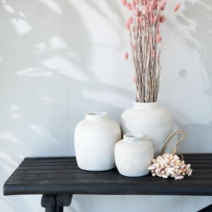 Drie witte vazen met roze gedroogde bloemen op zwart houten tafelblad, tegen een lichte muur.