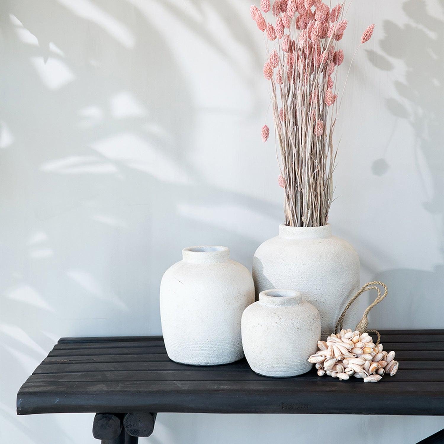 Rustieke witte vazen op houten plank, gevuld met roze gedroogde bloemen en schelpdecoratie ernaast.