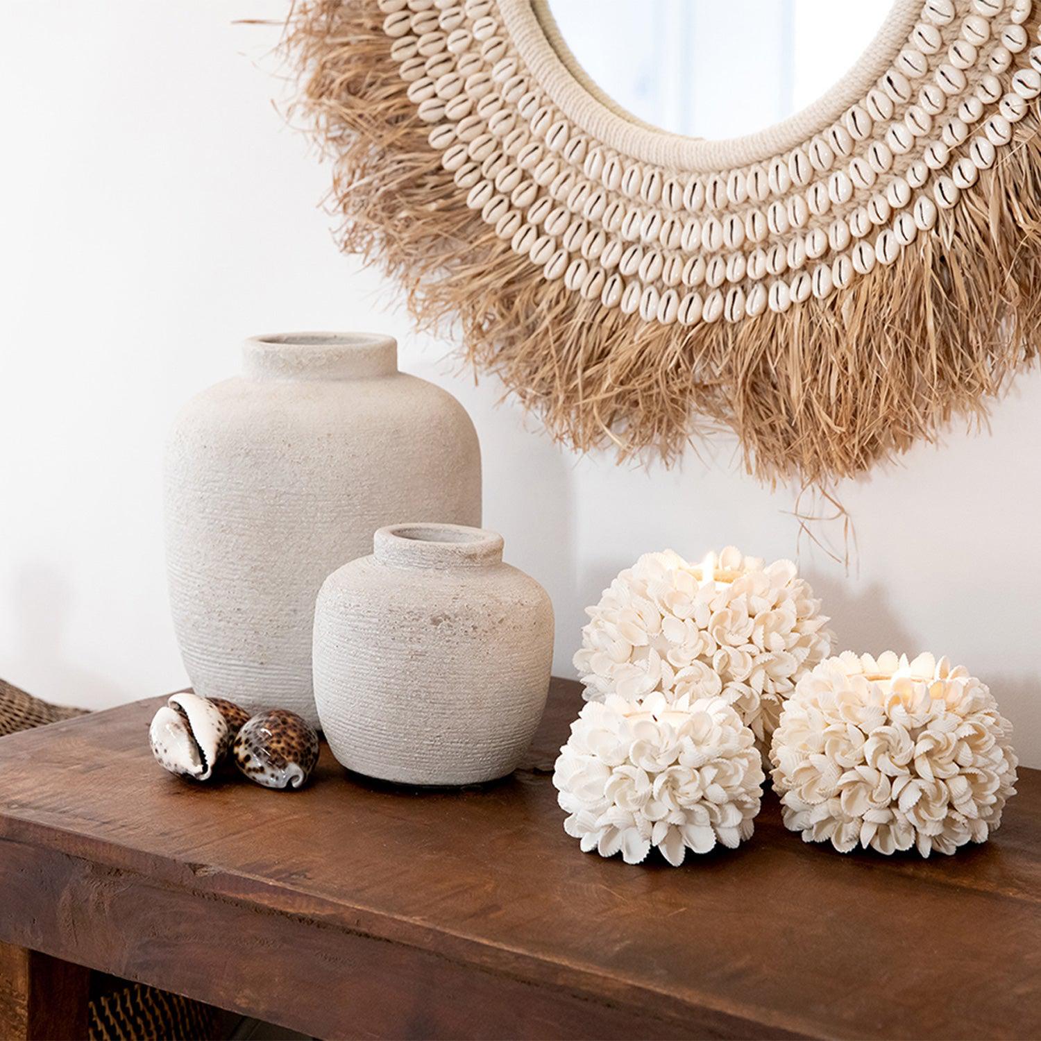 Witte keramische vazen en schelpenkaarsen op houten tafel, voor ronde spiegel met natuurlijke vezels.