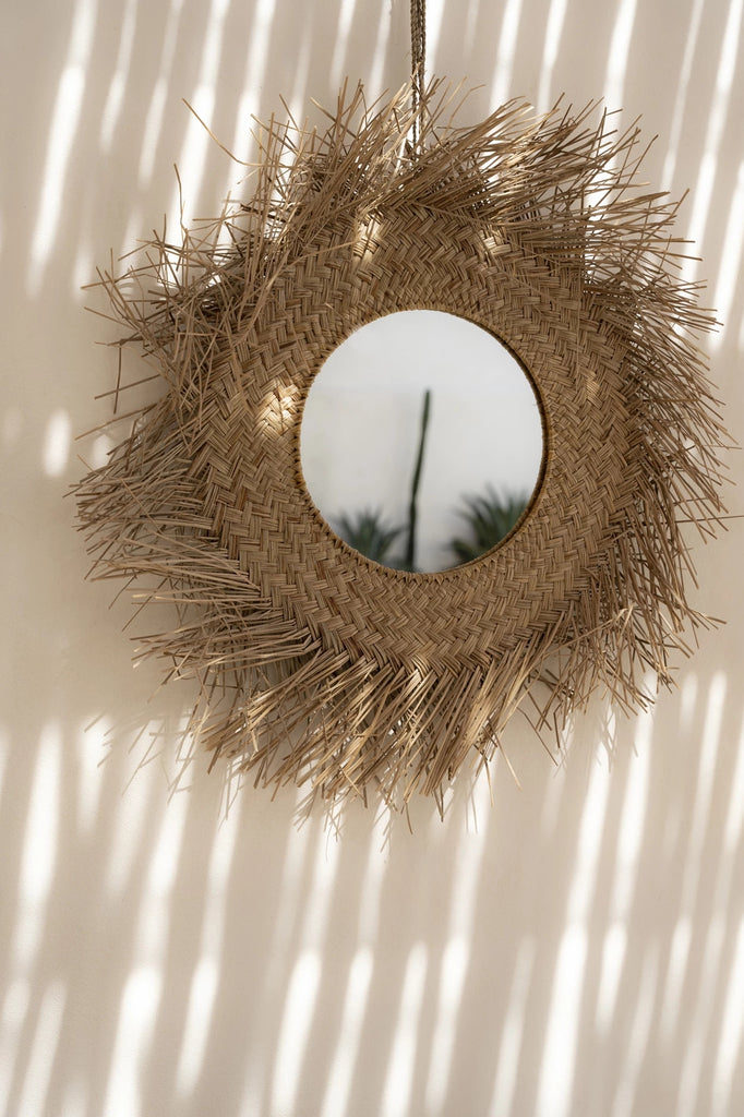 Ronde spiegel met een gevlochten rotan frame, omgeven door zonlicht en schaduwen op een witte muur.
