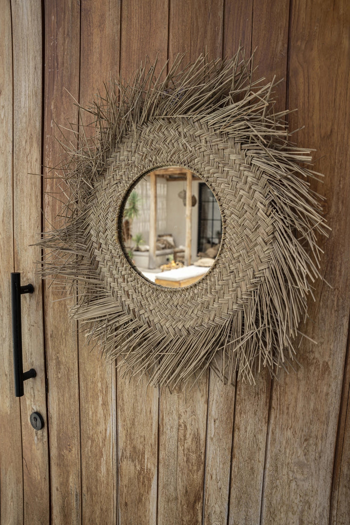 Rieten spiegel met een kunstzinnig patroon, reflecteert een rustieke binnenplaats op een houten deur.