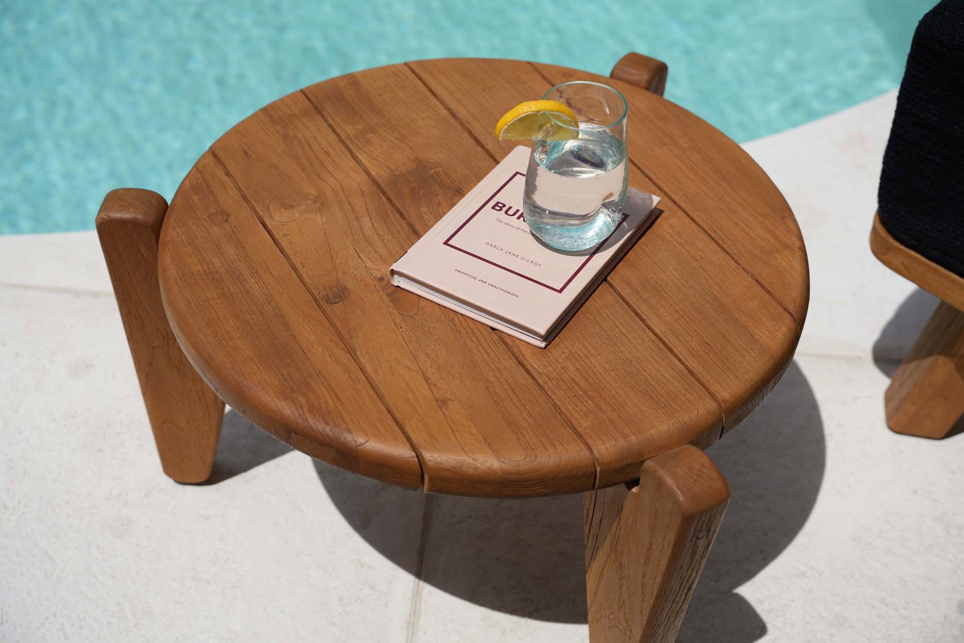 Close-up van ronde Seseh salontafel in gerecycled teak met glas water en hardcover boek op strak tegelterras, subtiel gestyled door Bazar Bizar