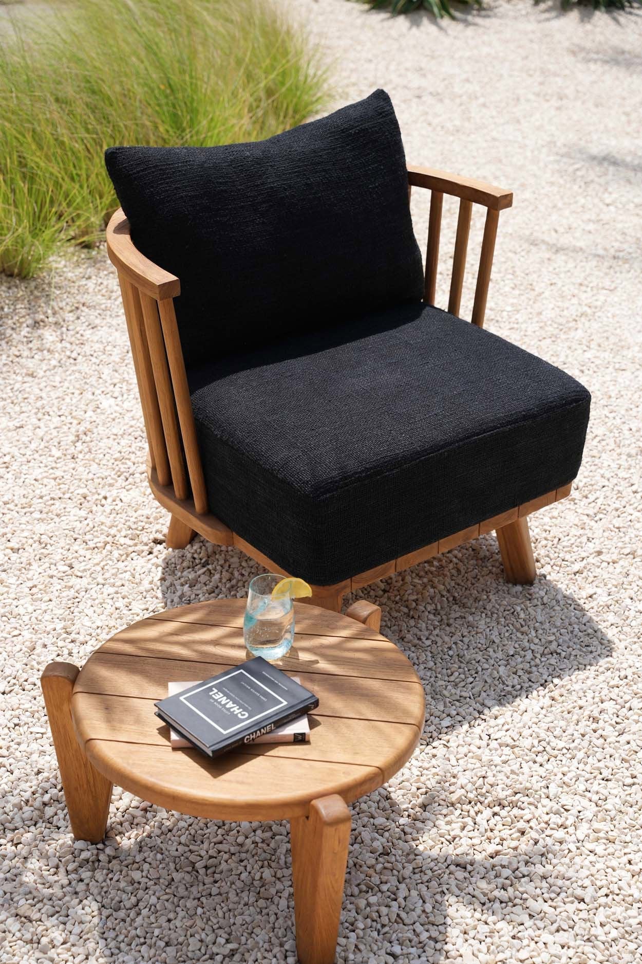 Warme naturel teakhouten Seseh salontafel van Bazar Bizar naast zwarte buitenfauteuil met kussen op wit grind