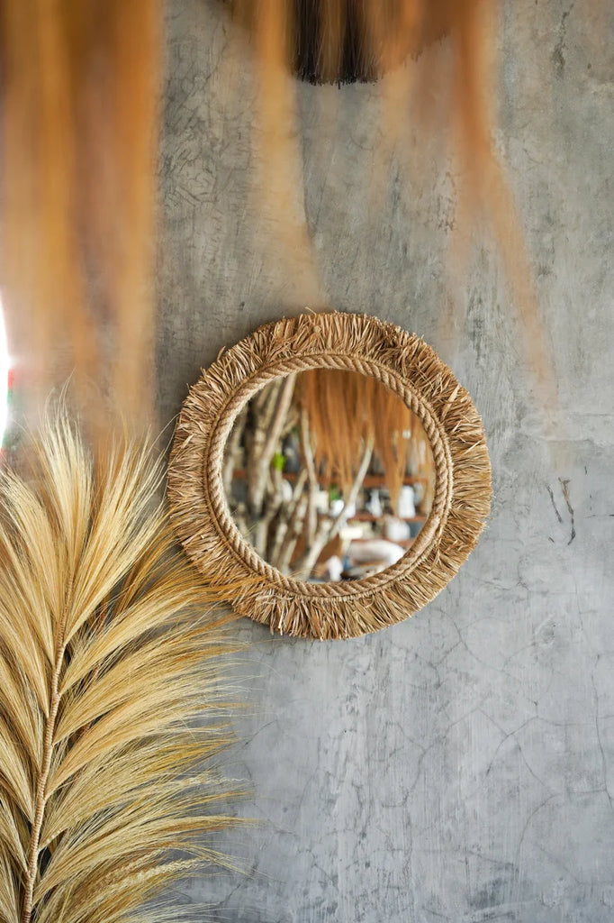 Ronde spiegel met natuurlijke rand reflecteert interieur, naast pluim van beige gras tegen ruwe, grijze muur.