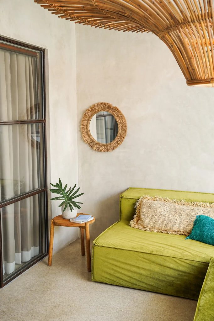 Olijfgroene bank met rotan spiegel aan muur, houten bijzettafel met plant en wit boek op beige vloer.
