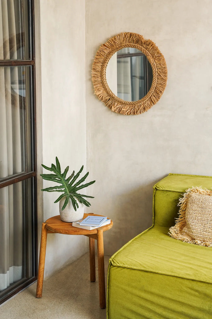 Houten bijzettafel met plant en boeken, groene fluwelen stoel en ronde, rustieke spiegel op beige muur.
