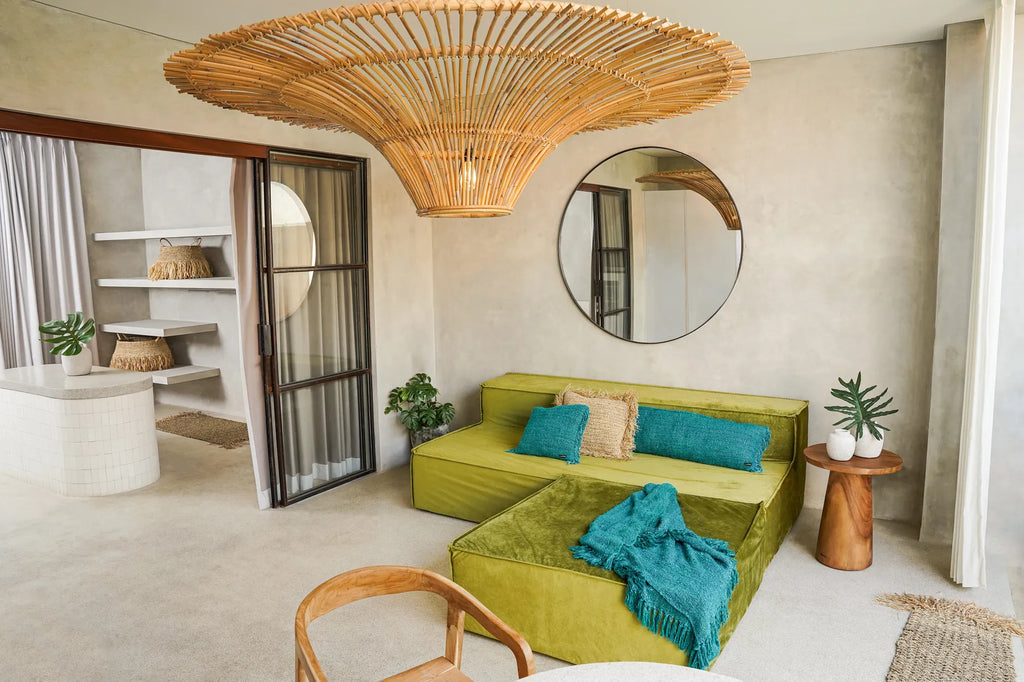 Stijlvolle woonkamer met groene fluwelen bank, blauwe kussens, rotan lamp en ronde spiegel op beige muur.