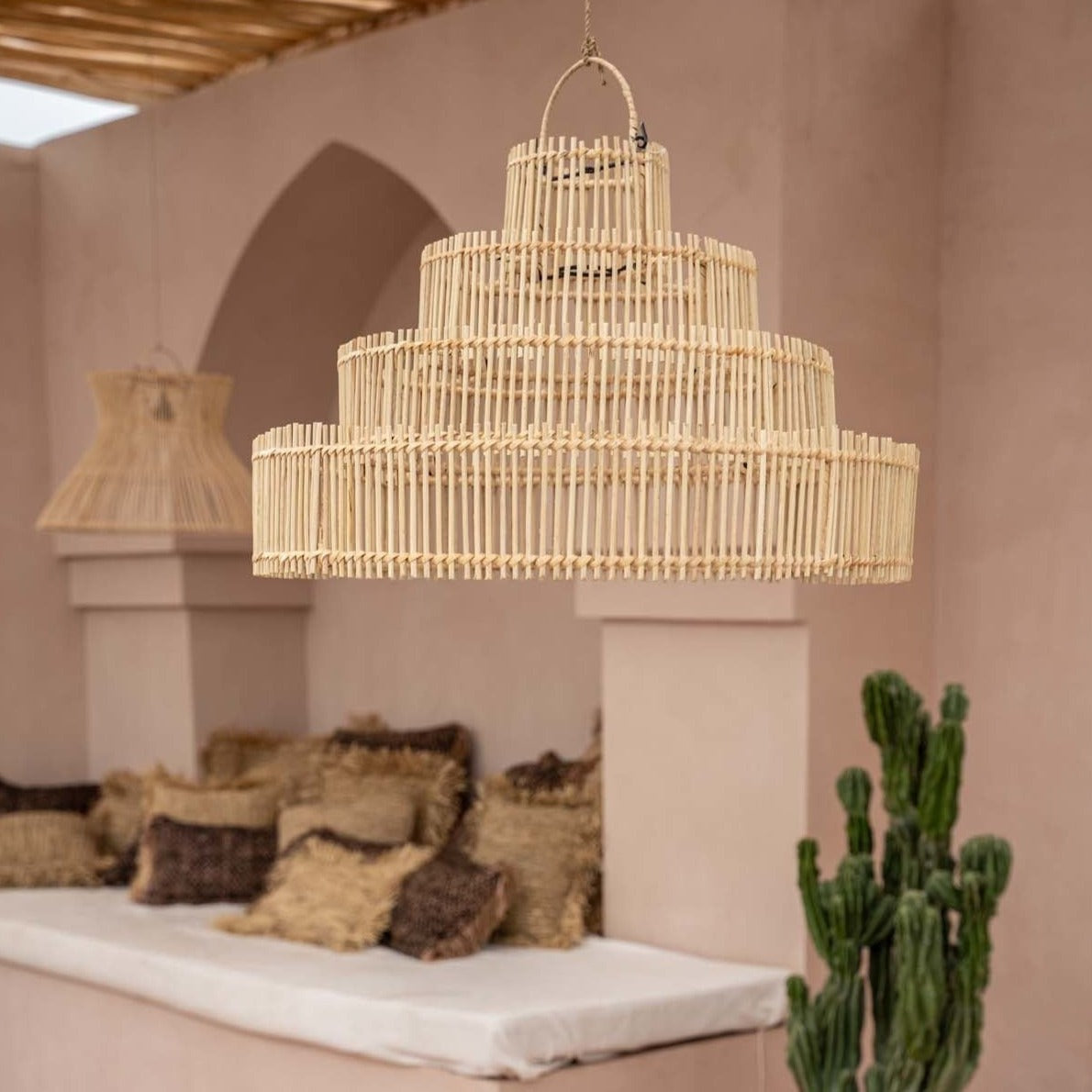 Grote, ronde bamboe hanglamp boven een beige zithoek met kussens en een groene cactus in een rustieke omgeving.