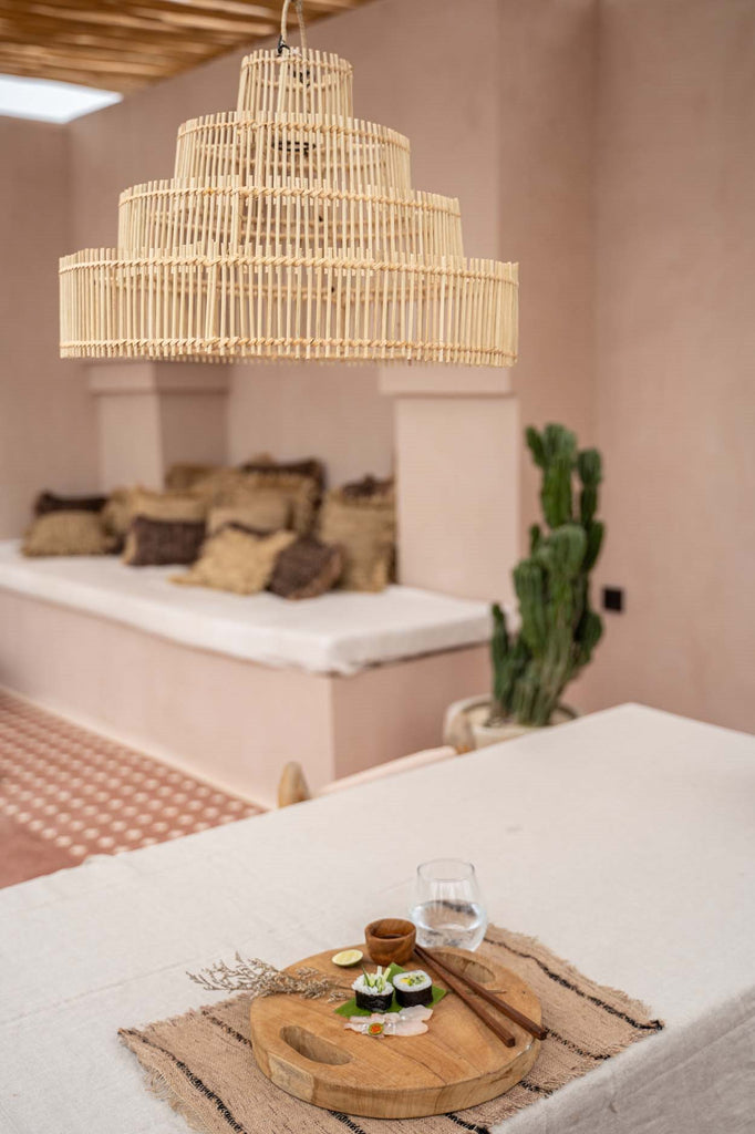 Houten plank met sushi en een glas water op een tafel, onder een hanglamp, met beige wanden en cactussen.