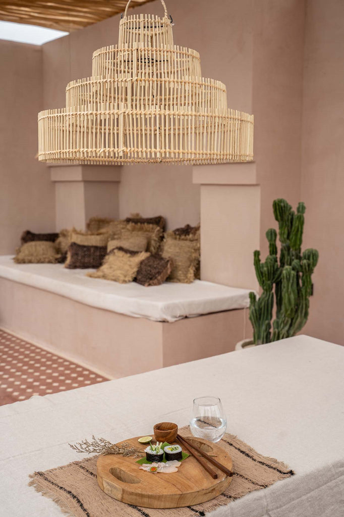 Rustieke eetkamer met grote rotan lamp, houten snijplank met sushi, waterglas en cactussen in de hoek.