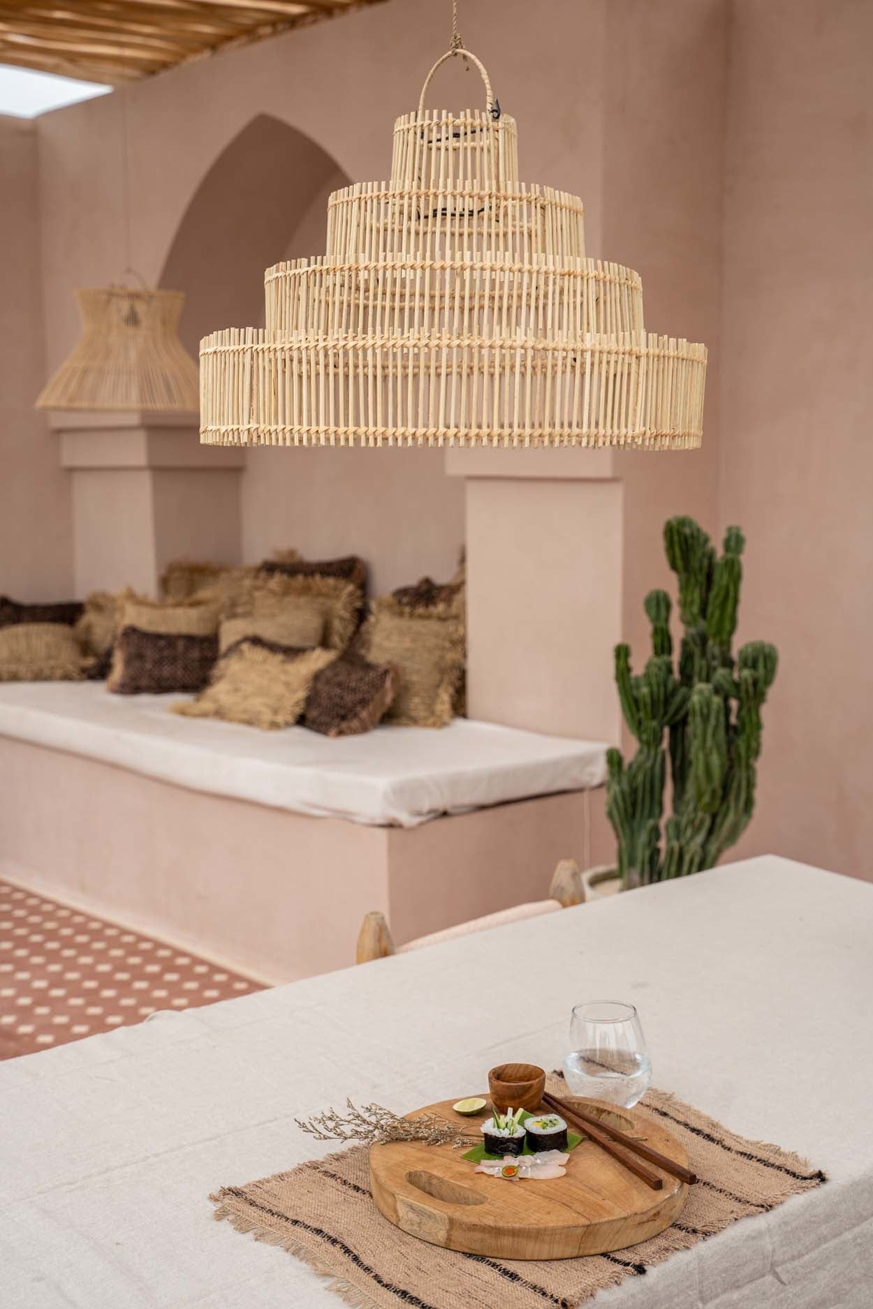 Houten loungebank met kussens, bamboe hanglamp, houten schaal met sushi op tafel en een cactus op achtergrond.
