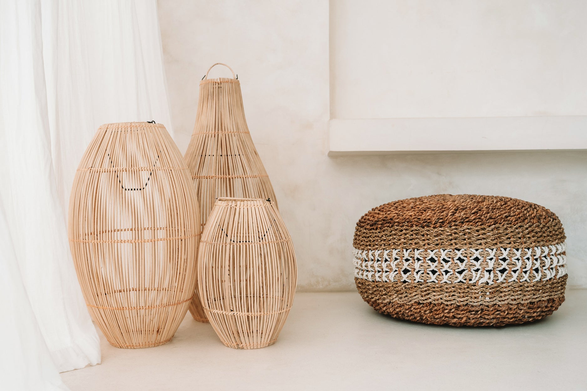 Drie cilindrische rotan lampen naast een ronde, gevlochten poef met witte decoratie op beige vloer.