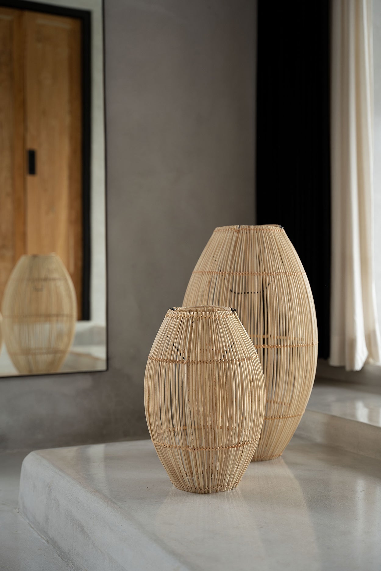 Twee elegante, geweven houten vazen op een minimalistisch betonnen plateau naast een grote spiegel.
