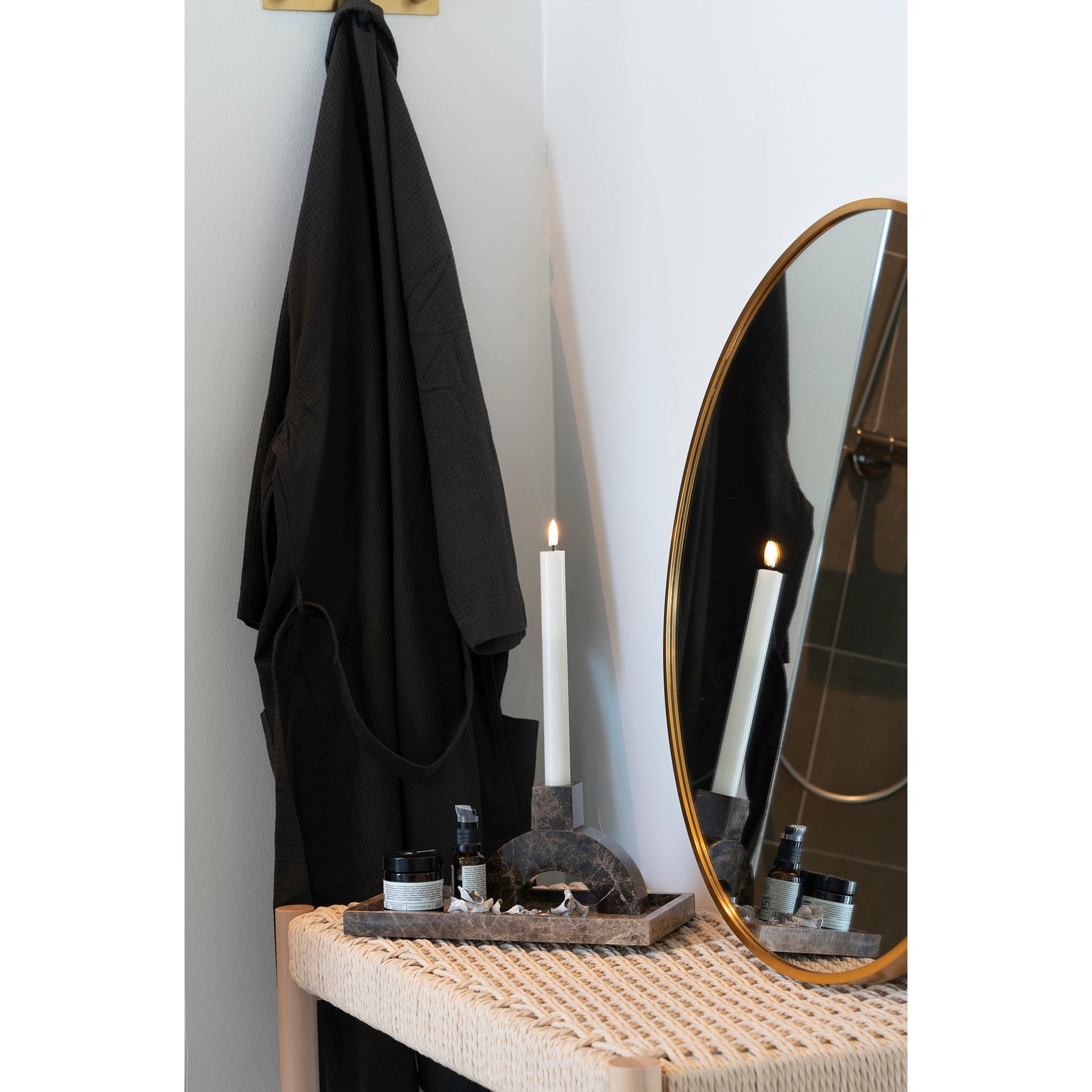 Zwarte badjas hangt naast tafel met spiegel, brandende kaars en verzorgingsproducten in zen-sfeer badkamer.