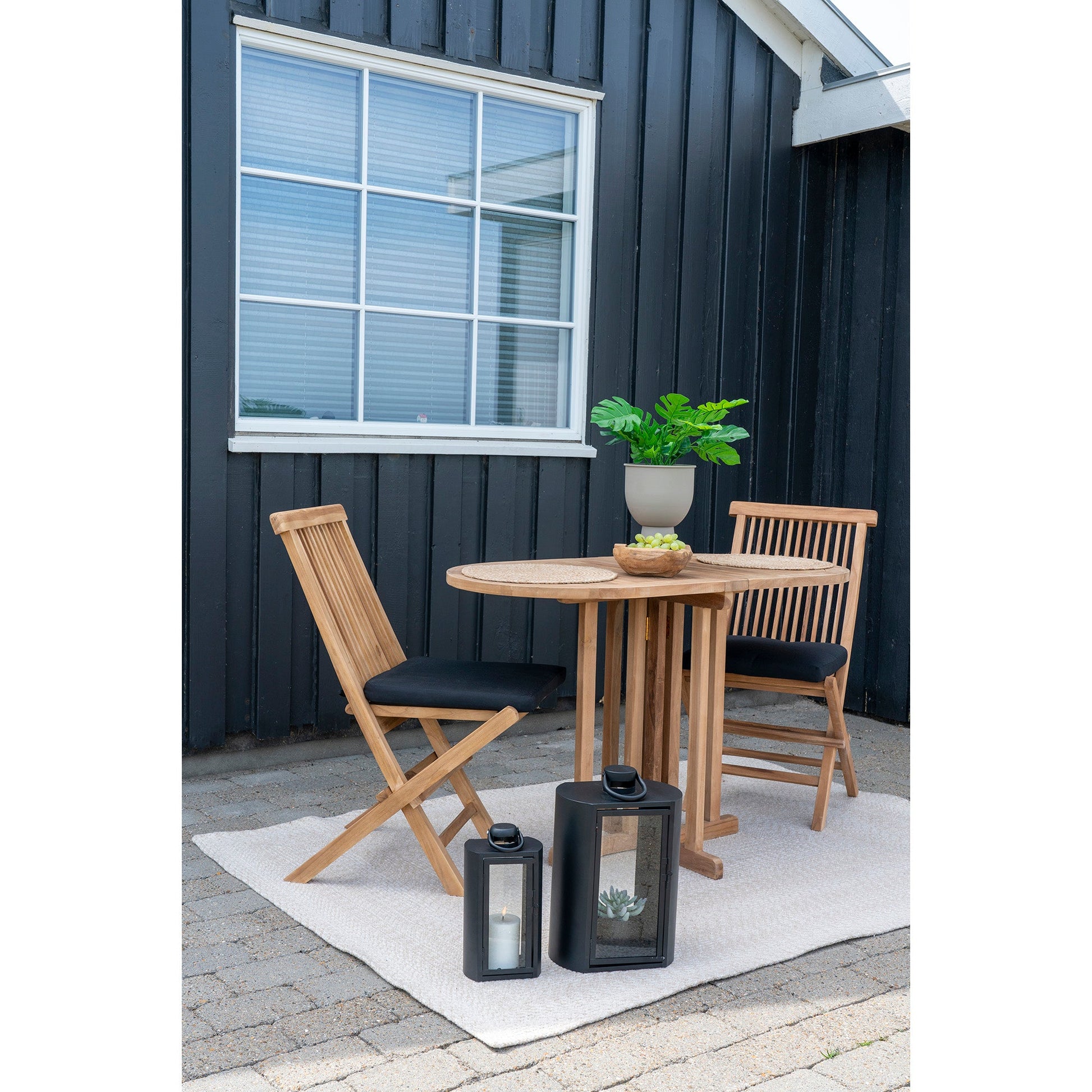 Houten tuinset met ronde tafel, groene plant en fruitschaal. Zwarte lantaarns op tapijt, tegen donker houten gebouw.