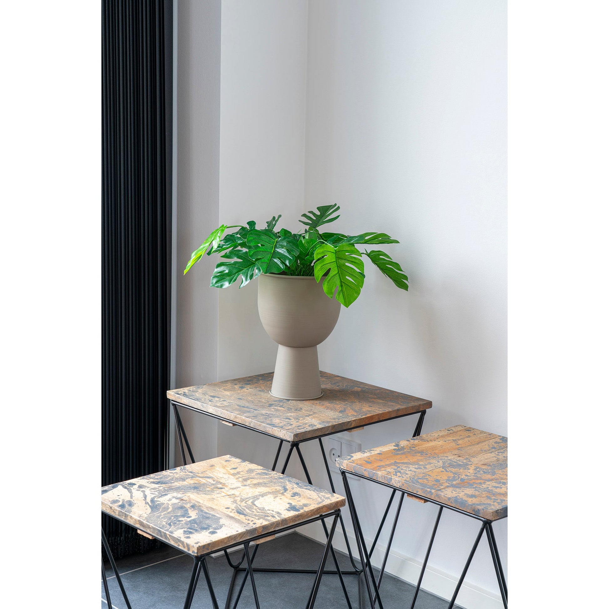 Grijze vaas met groene Monstera plant op rustieke houten tafels met metalen poten in een lichte kamer.