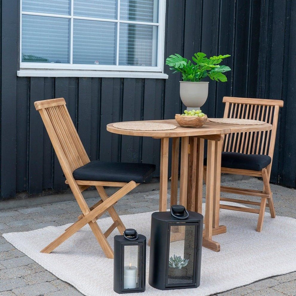 Houten tafel en stoelen met zwarte kussens, groene plant, schaal met druiven op een lichte mat voor donkere muur.