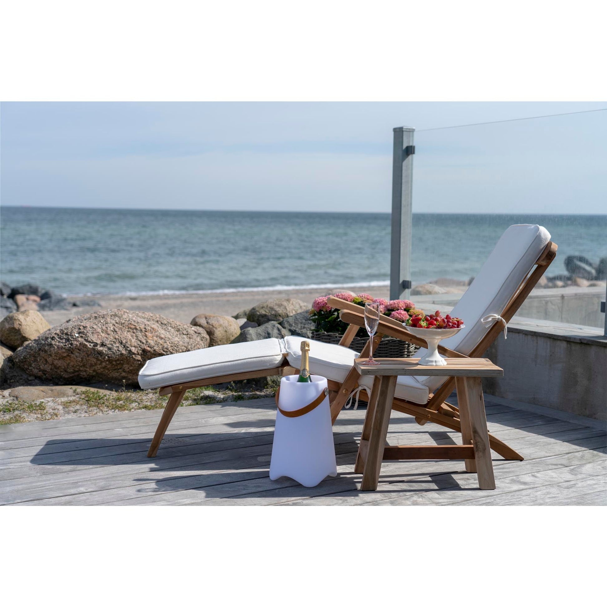 Luxe ligstoel met kussens op terras, uitzicht op zee, champagne in koeler, schotel met bessen en bloemen.