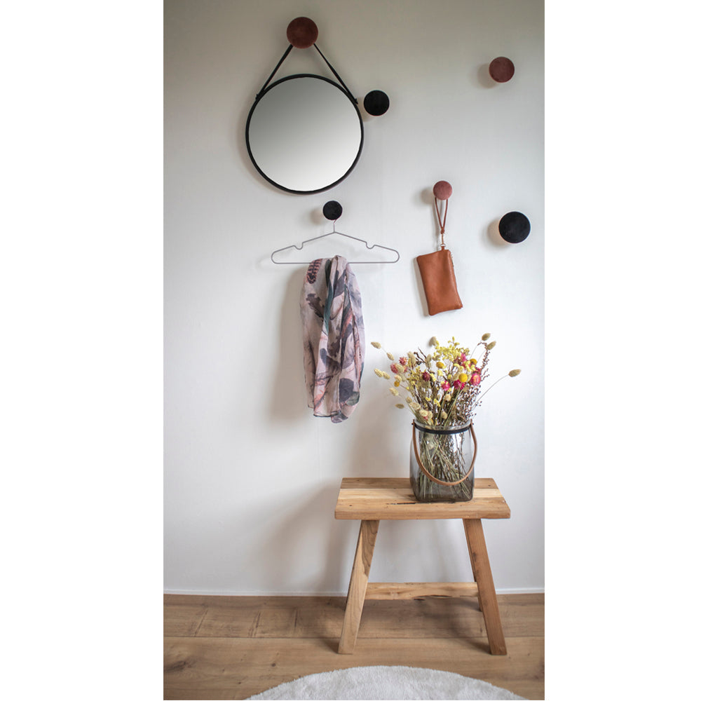 Rond spiegel met leren riem, bloemenvaas op houten bankje en sjaal hangen aan muur met bruine knoppen.