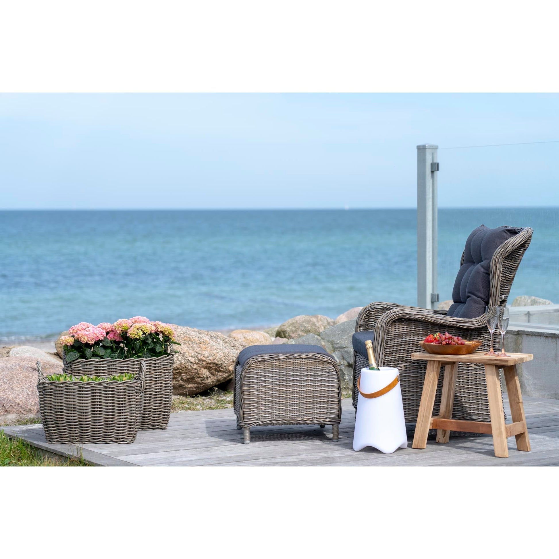 Rotan stoel en voetenbank op terras aan zee, naast tafel met fruitschaal en champagnekoeler, bloemen op achtergrond.
