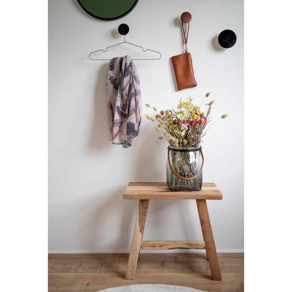 Houten bankje met glazen vaas vol kleurrijke droogbloemen, bruine tas en sjaal aan witte muur.