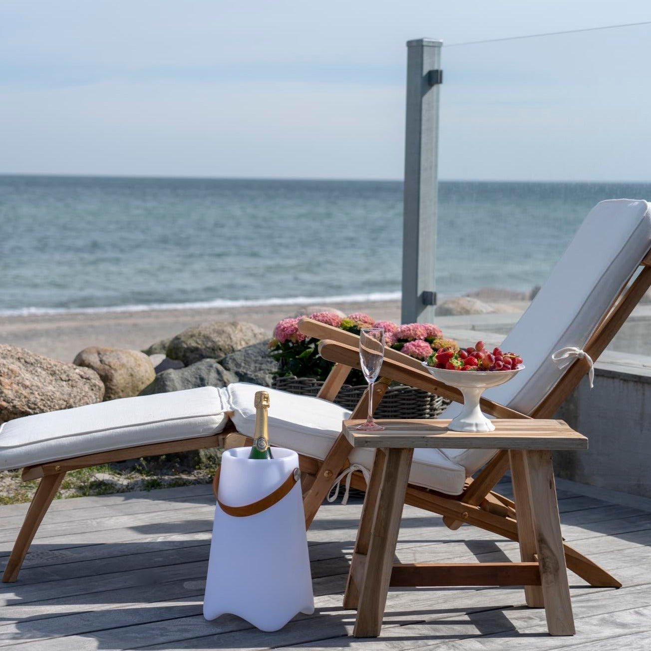 Ligstoel met wit kussen, houten bijzettafel met champagne en schaal druiven, zee in de achtergrond.