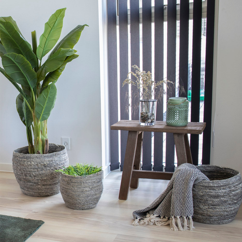 Banenenplant in een rieten pot, naast een houten bank met glazen vaas en groene kaars, naast zwarte lamellen.