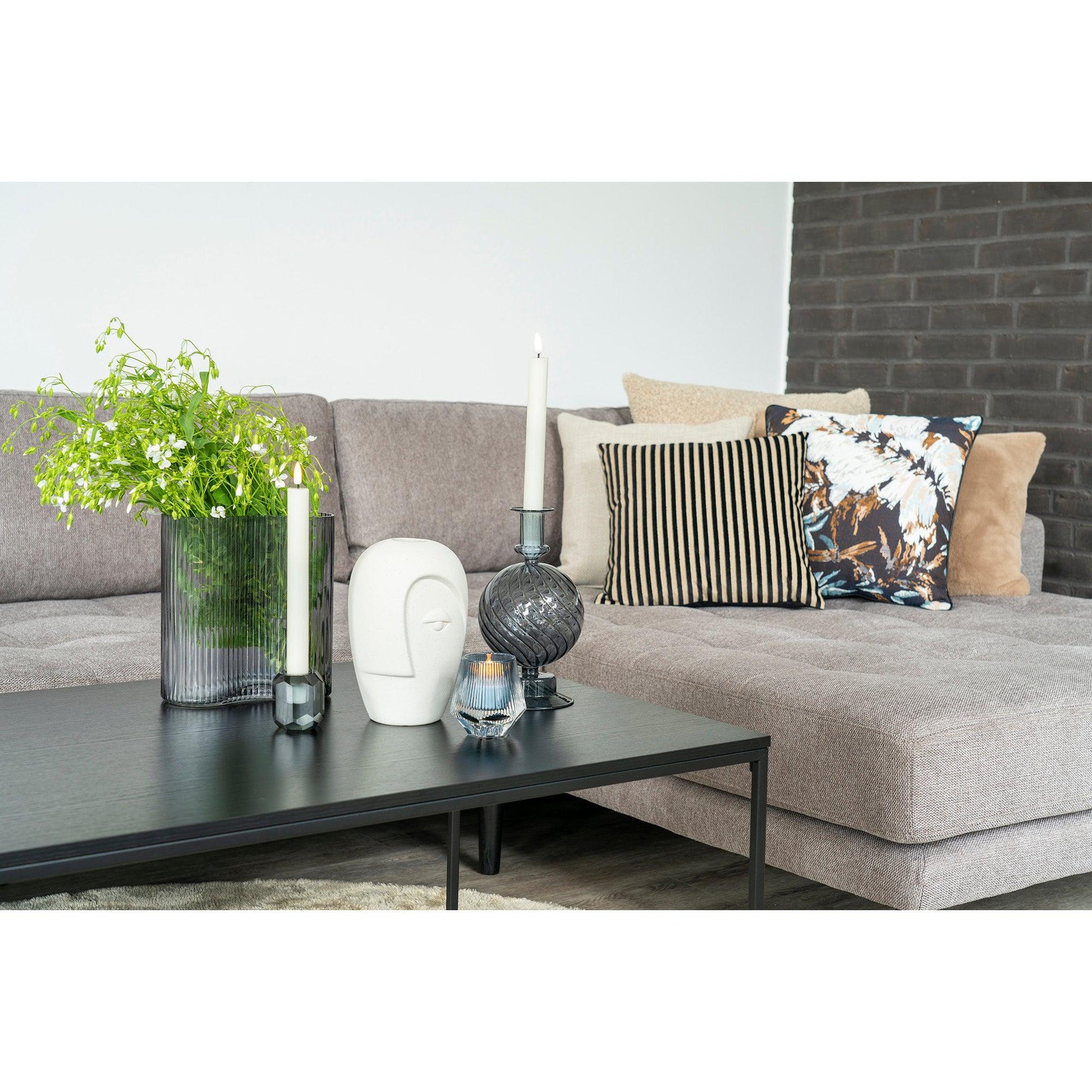 Grijze bank met decoratieve kussens, moderne vaas met groene plant, en kaarsen op zwarte salontafel.