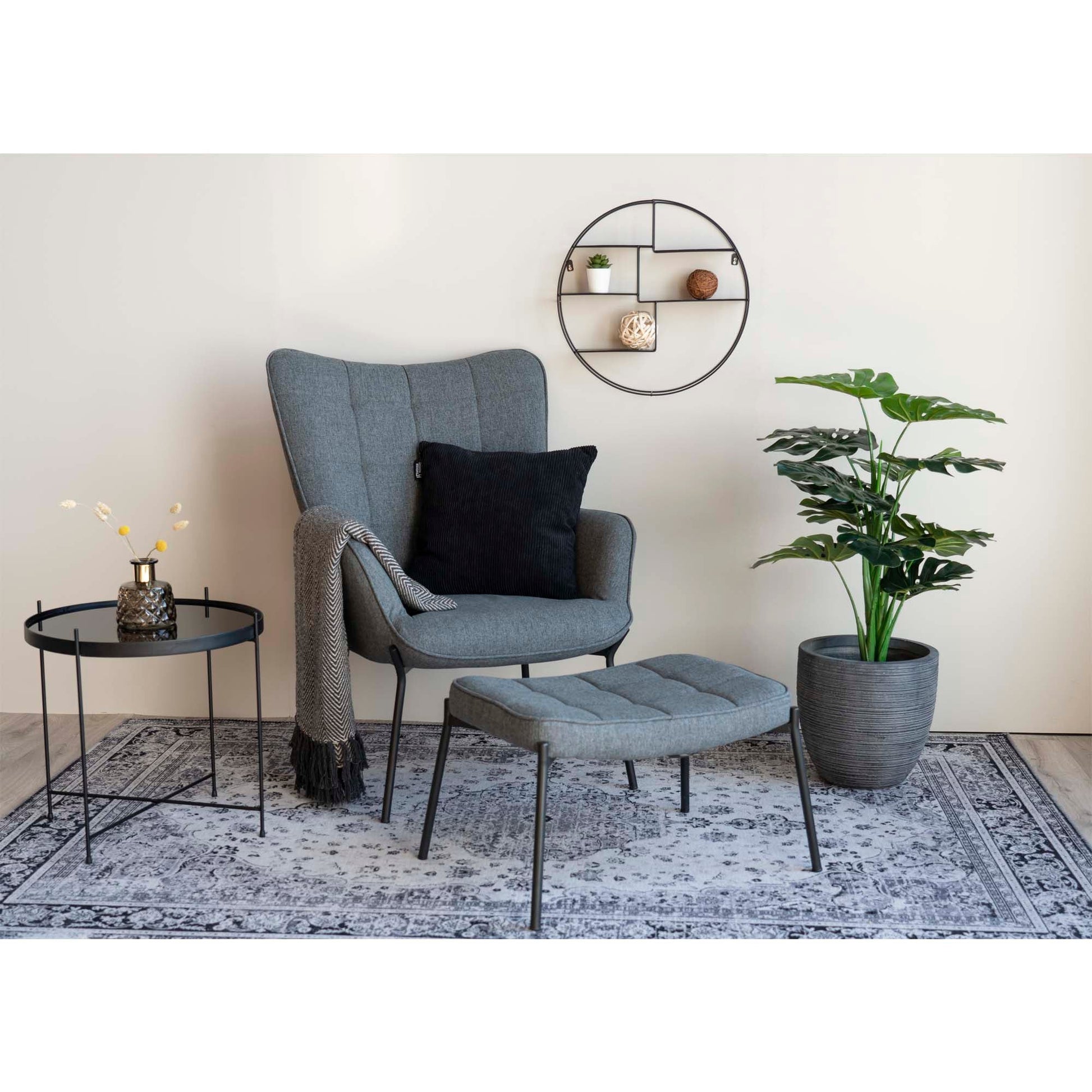Grijze fauteuil met zwart kussen, poef en bijzettafel op tapijt, naast een grote groene plant en wandrek.