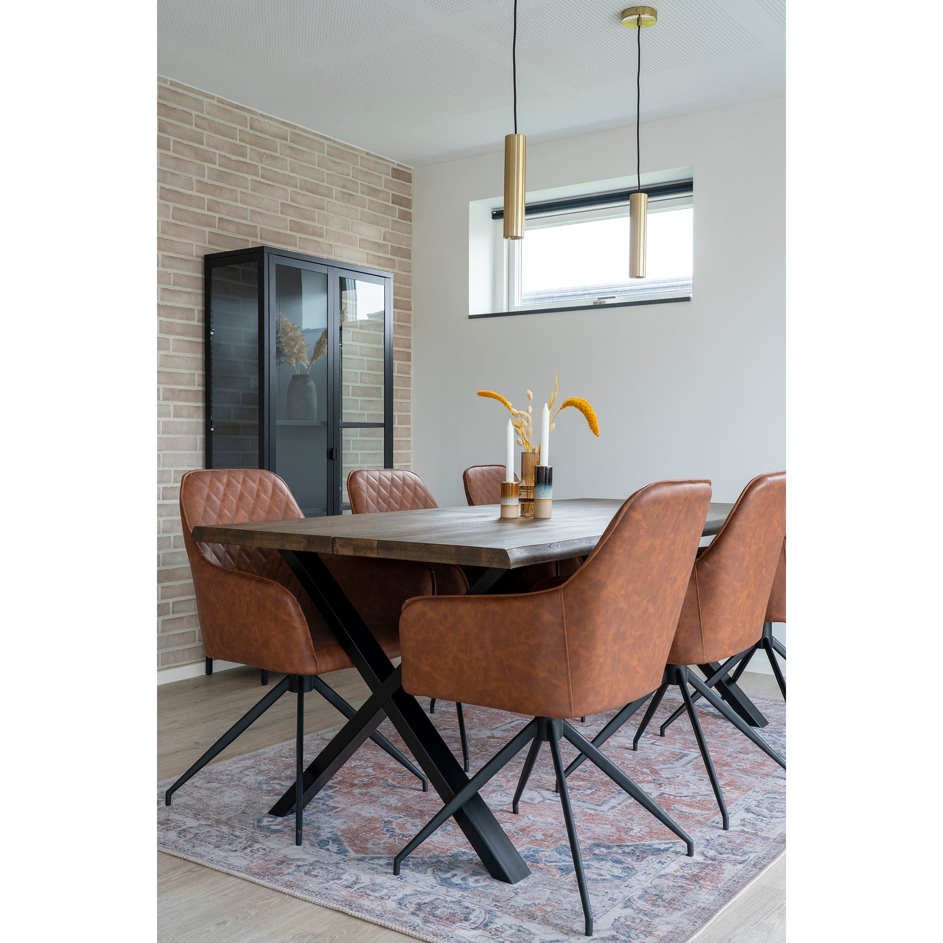 Bruine leren stoelen rond houten tafel op tapijt, gouden hanglampen, bakstenen muur, lichte en moderne sfeer.