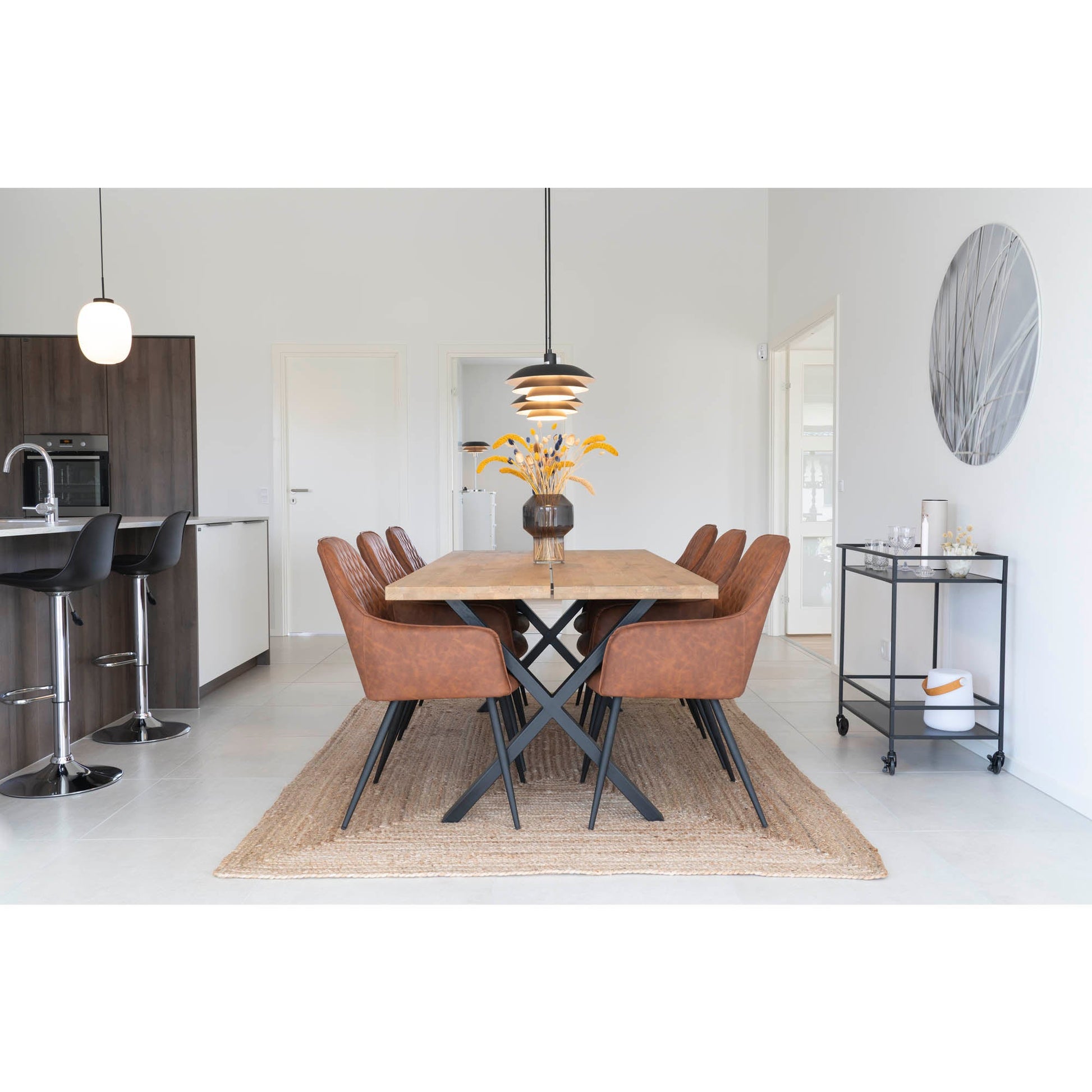 Moderne eetkamer met houten tafel, zes bruine leren stoelen, hanglamp en vaas met gele bloemen op sisal tapijt.