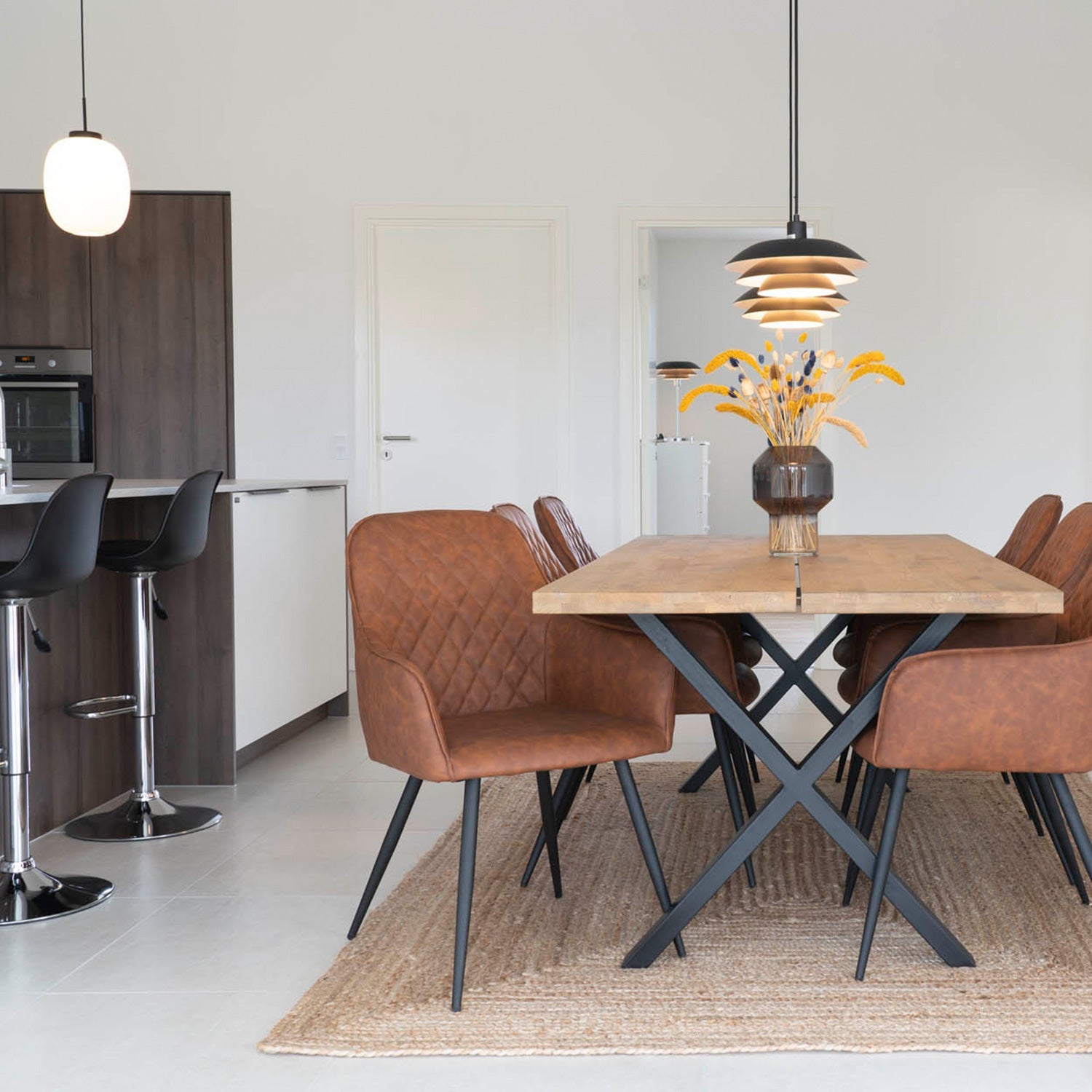 Eettafel met bruine leren stoelen, houten blad, metalen poten, zwarte hanglamp en vaas met droogbloemen.
