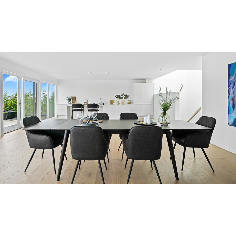 Strakke eetkamer met zwarte stoelen, een lange tafel en vaas met groen decor; grote ramen geven uitzicht.