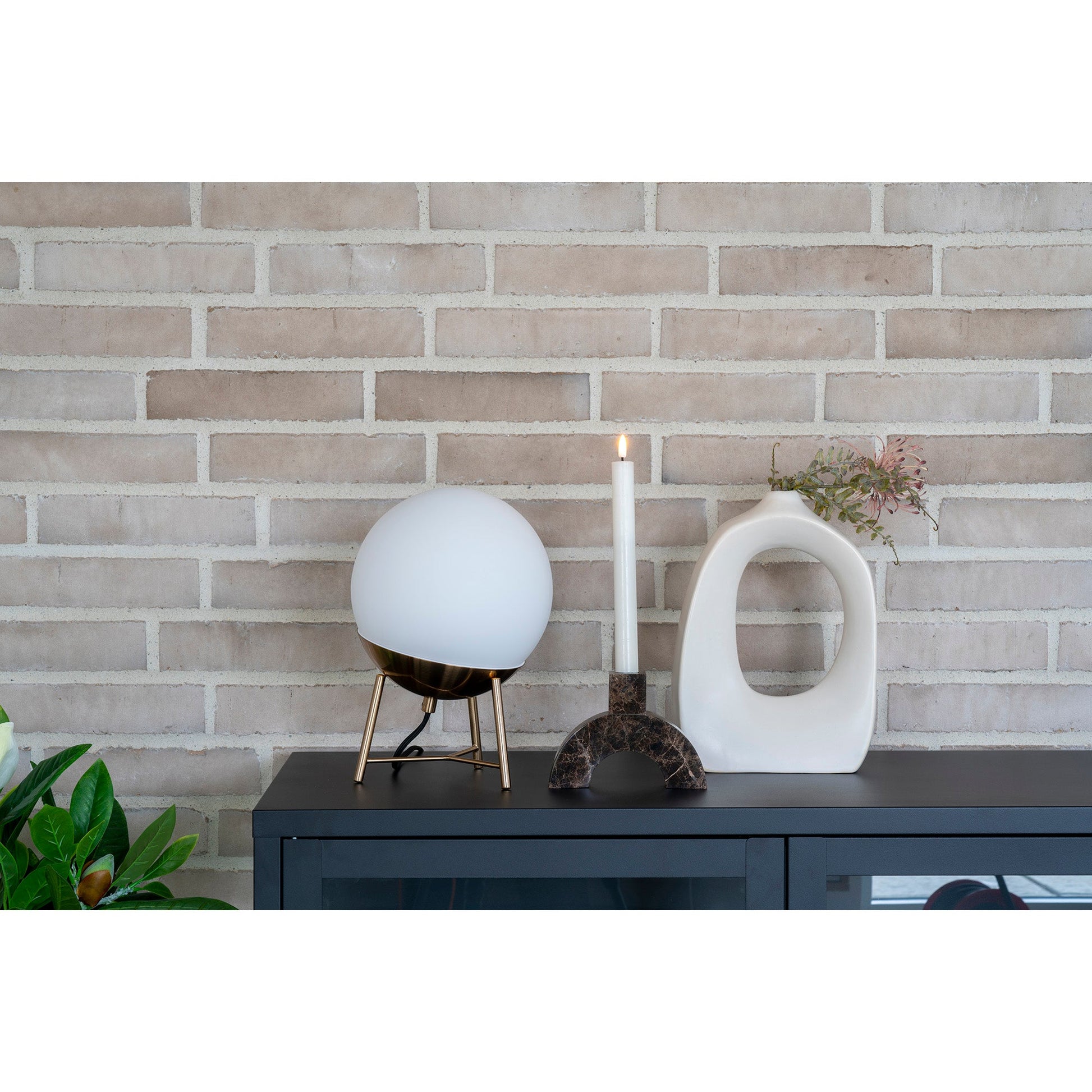 Moderne opstelling met witte bolvormige lamp, brandende kaars en keramische vaas tegen bakstenen wand.