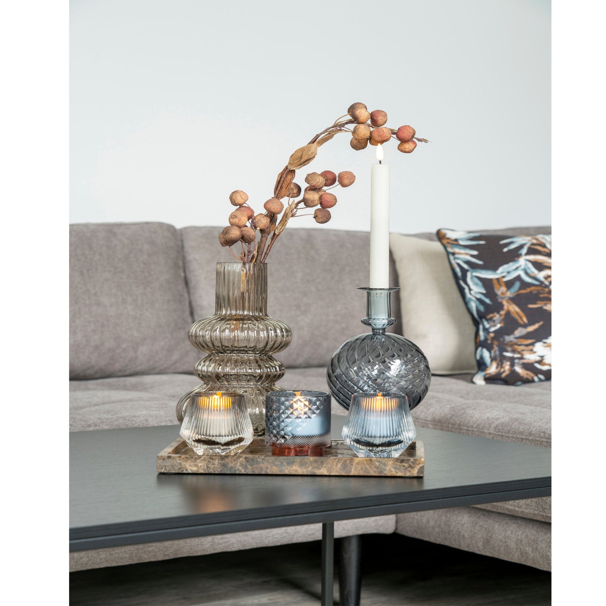Glazen vazen met gedroogde bloemen en kaarsen op een houten plateau, voor een beige bank met sierkussen.