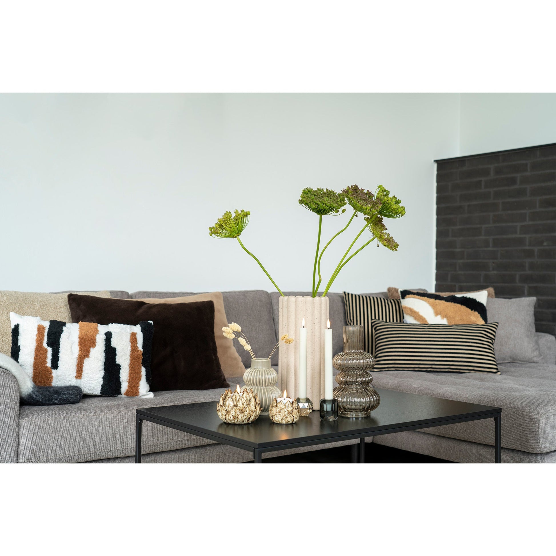 Grijze bank met diverse kussens; zwarte salontafel met vaas, kaarsen en decoratieve bloemen. Sfeervol interieur.