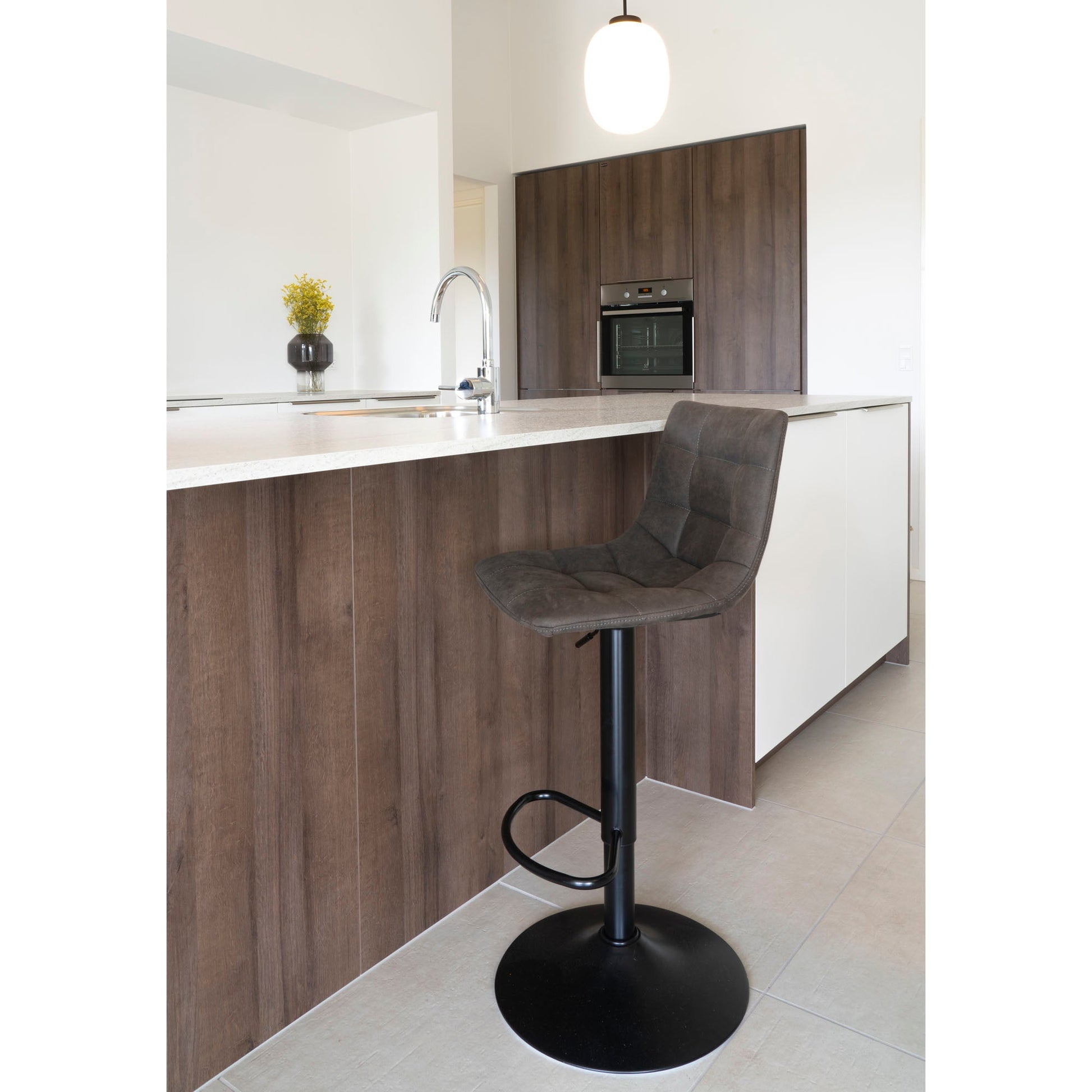 Moderne keuken met houten kastdeuren, wit aanrecht en grijze barkruk. Hanglamp en vaas met gele bloemen.