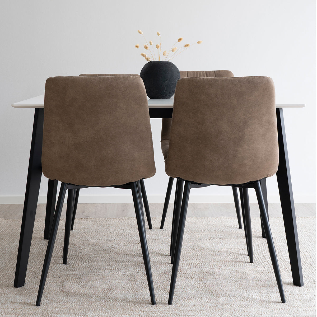 Vier bruine suede eetkamerstoelen bij een zwarte tafel, gedecoreerd met een zwarte vaas met gedroogde bloemen.