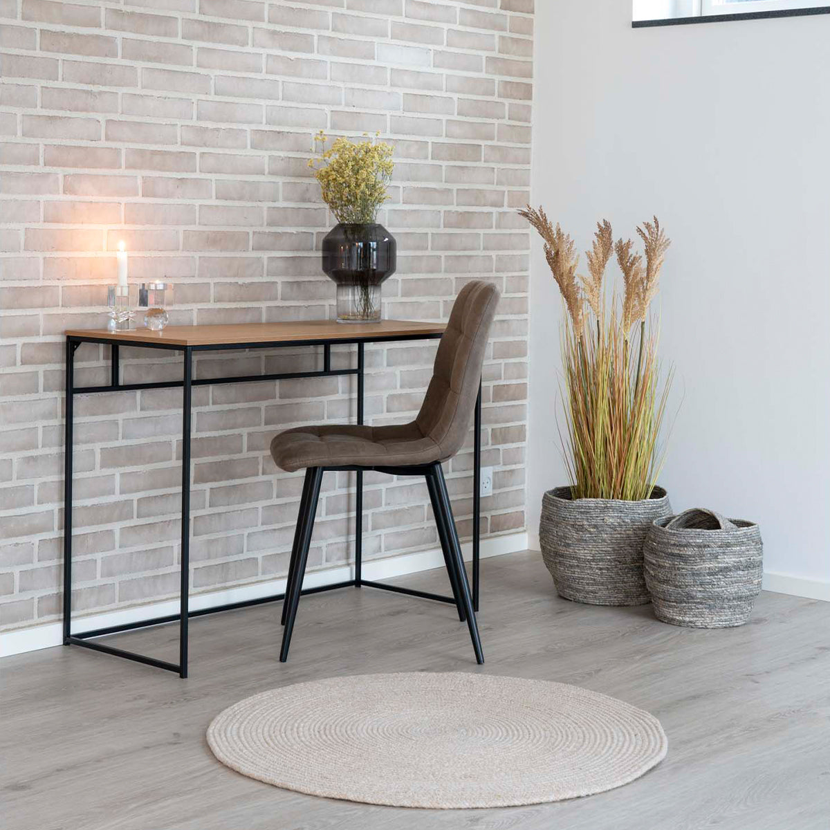 Stijlvolle werkhoek met houten bureau, bruine stoel, vaas met bloemen en sfeervolle kaars tegen bakstenen muur.