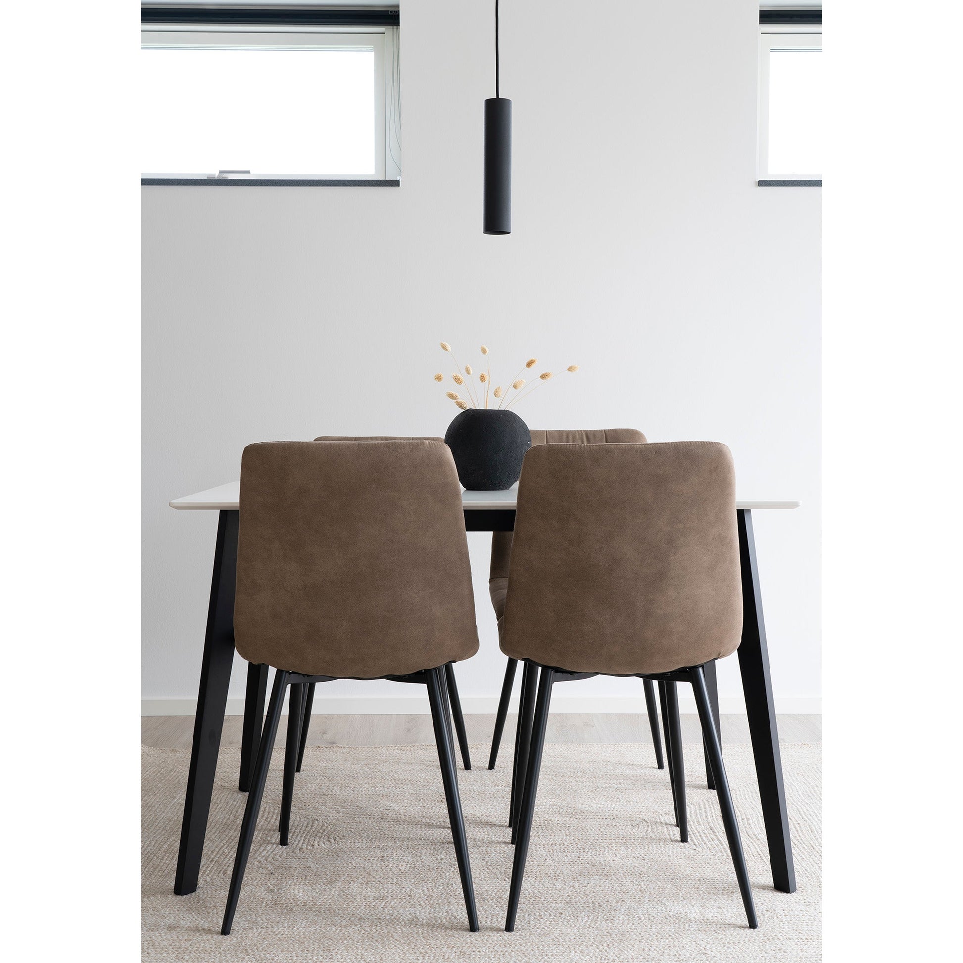 Eenvoudige eetkamer met bruine stoelen, zwarte vaas met droogbloemen en een hanglamp tegen witte muren.