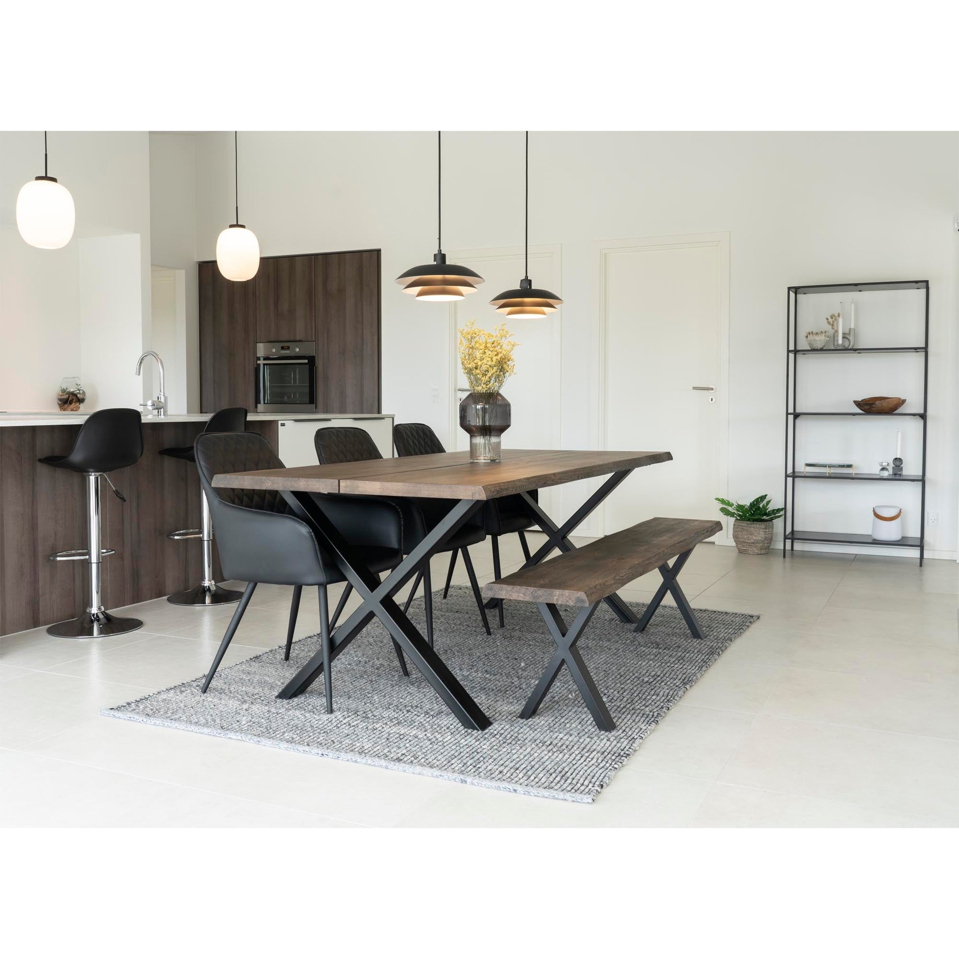 Moderne eetkamer met houten tafel, zwarte stoelen en bank, hanglampen en witte keuken op achtergrond.