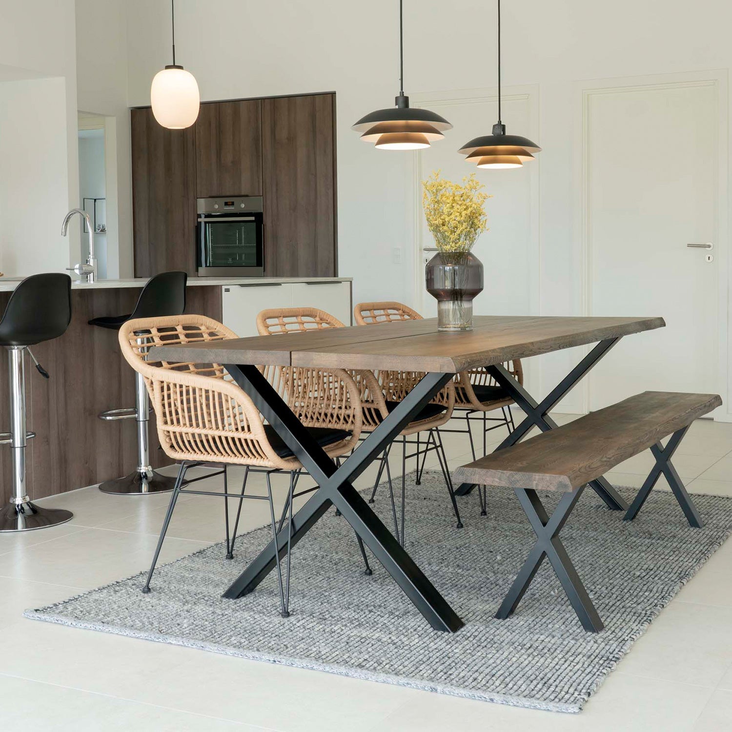 Modern eetkamer met houten tafel en banken, rieten stoelen, en bovenhangende lampen in lichte, ruime keuken.