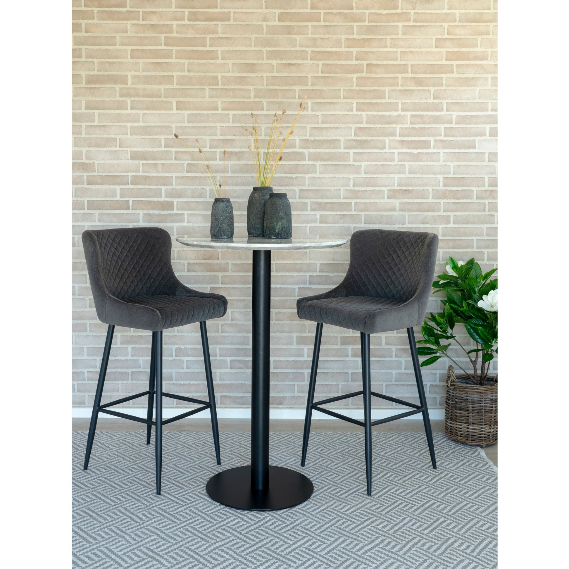 Twee grijze barkrukken en een ronde tafel met vazen tegen beige metselwerk, naast een plantmand.