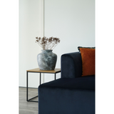 Vita bijzettafel van House Nordic naast blauwe sofaarm met kussens, met gouden tafellamp en amberkleurige vaas.