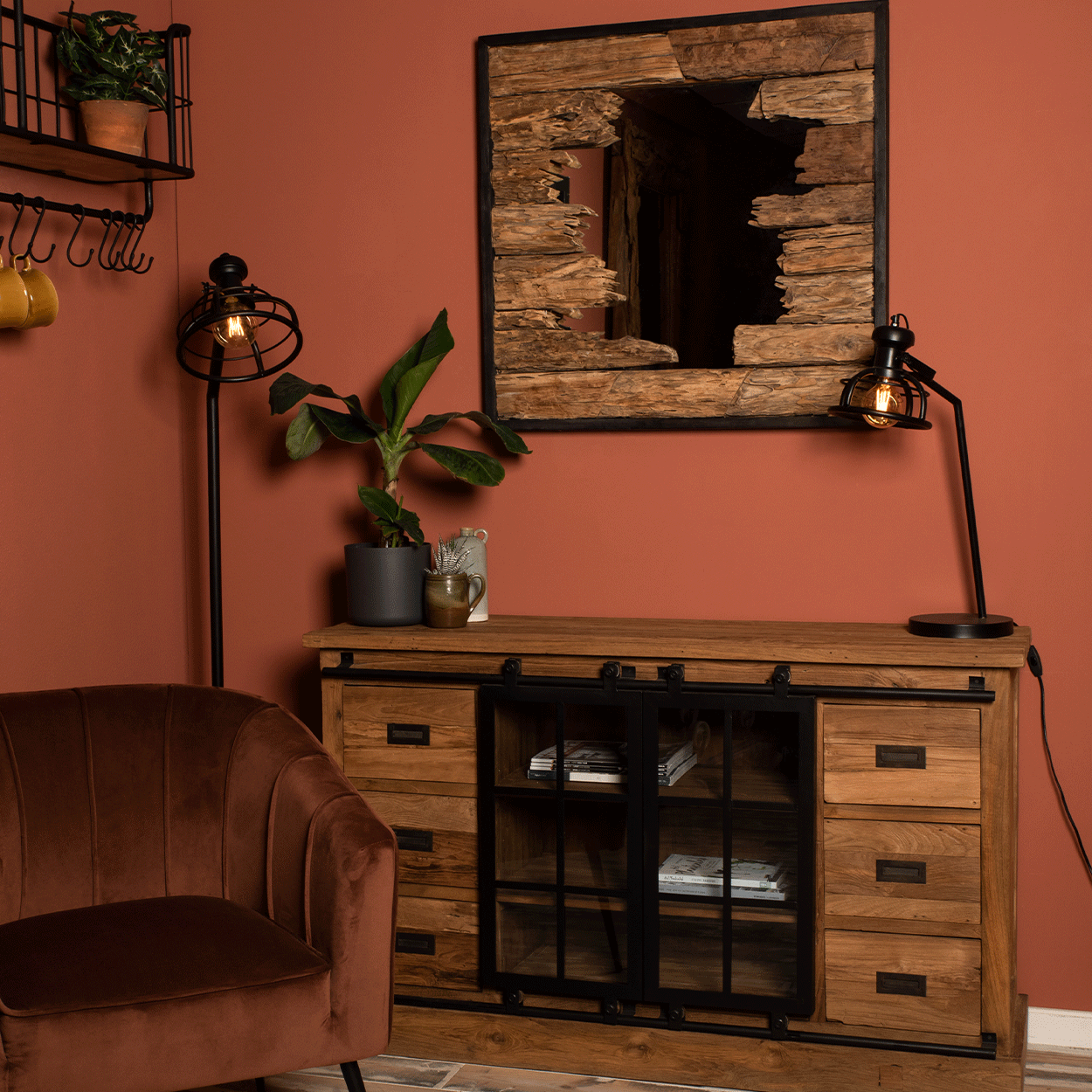 Rustieke kamer met houten commode, spiegel en bruine stoel tegen terracotta muur. Planten en lampen sieren de ruimte.