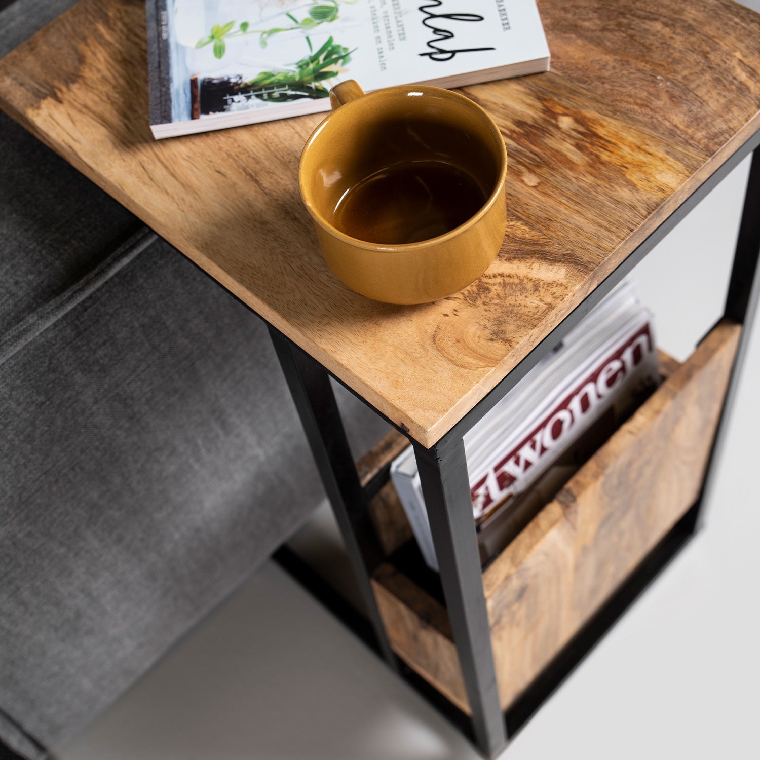 Gele mok met koffie op houten bijzettafel naast sofa; tijdschriftenrek met "Wonen" magazine.