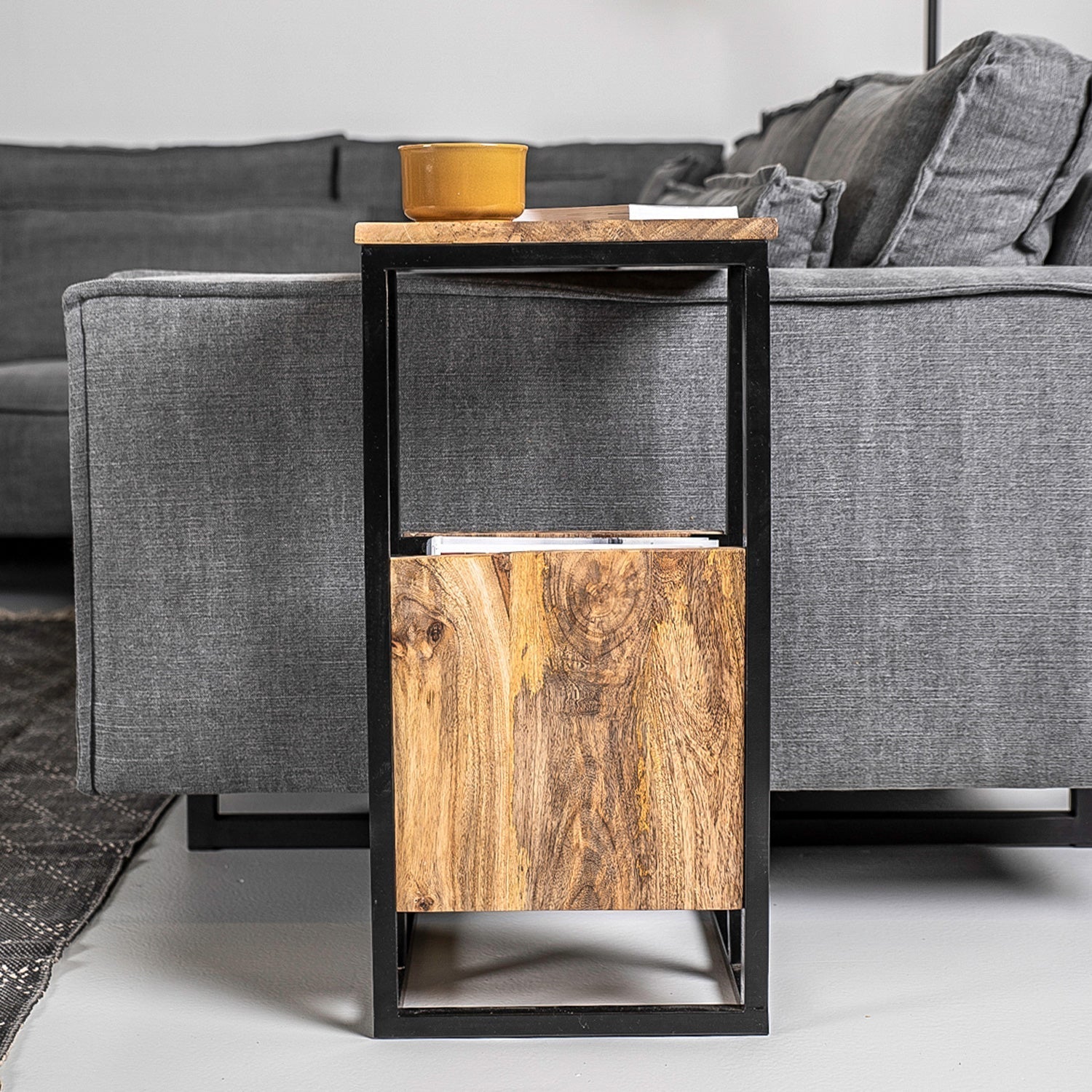 Strakke grijze bank met houten bijzettafel, metalen frame en mosterdgeel kopje bovenop, in moderne ruimte.