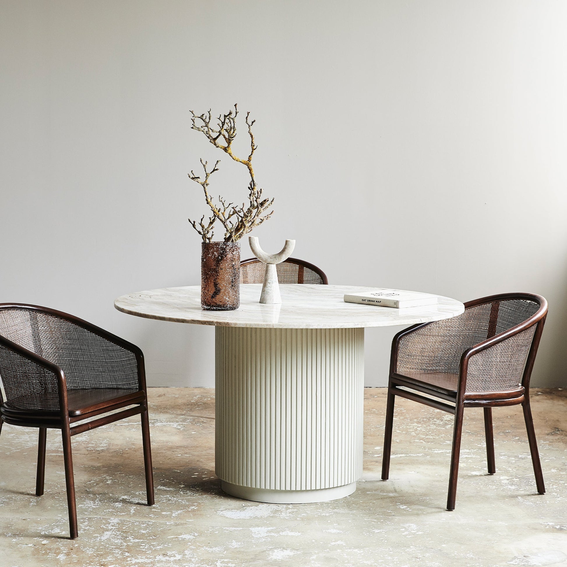 Ronde marmeren tafel met twee stoelen, decoratieve takken in vaas, en boek op een betonnen vloer.
