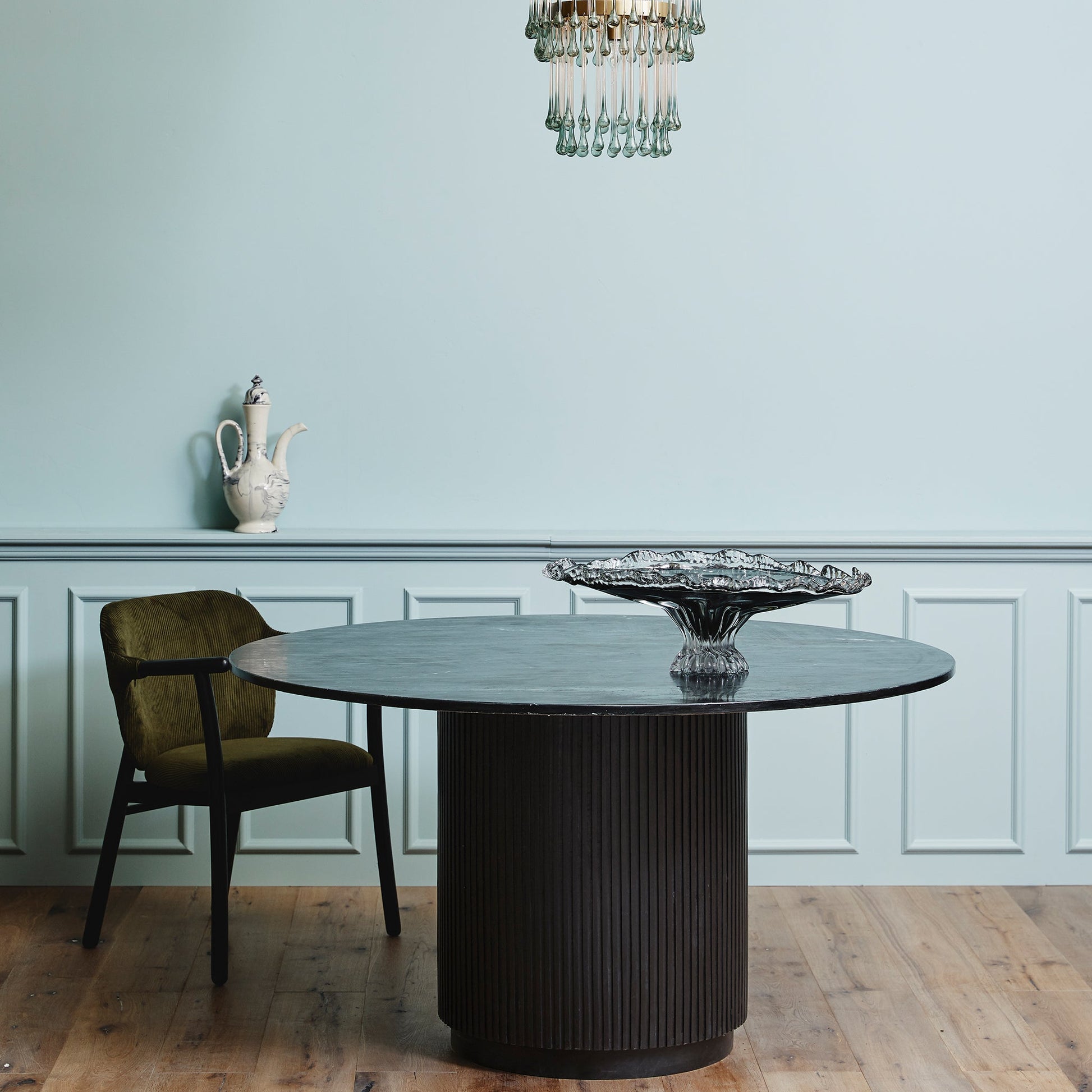 Ronde zwarte tafel met glazen schaal, groene stoel en sierlijke vaas tegen een lichtblauwe muur met kroonluchter.