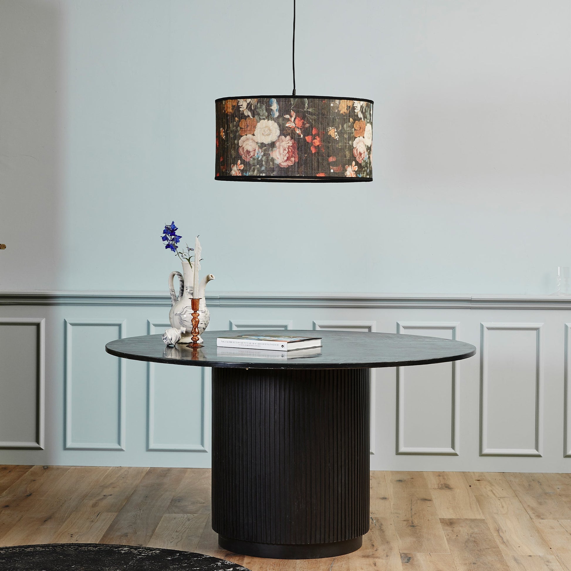Houten tafel met bloemenlamp en witte vaas met blauwe bloemen voor een lichtblauwe muur met lambrisering.